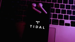 Yanında kablosuz kulaklık bulunan bir dizüstü bilgisayarın klavyesinde Tidal logolu akıllı telefon