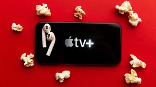 Apple TV Plus logolu iPhone, üstünde bir çift AirPods, patlamış mısır parçalarıyla kırmızı bir arka plan üzerinde duruyor