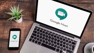 Ahşap bir masanın üzerinde duran bir dizüstü bilgisayarda ve telefonda görüntülenen Google Meet logosu