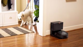 Köpekli bir adam odaya girerken iRobot Roomba J7+ koridordaki şarj istasyonunda