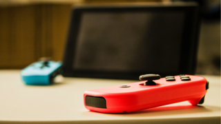 Ön planda bir masa üzerinde neon kırmızısı bir Joy-Con, bir Nintendo Switch ekranı ve arka planda masanın üzerinde duran mavi Joy-Con, biraz odak dışı