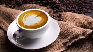 Bir kendir çuvalı ve kahve çekirdeklerinin yanında beyaz bir kahve fincanı ve tabağında üstte geç dönem sanatı olan bir espresso